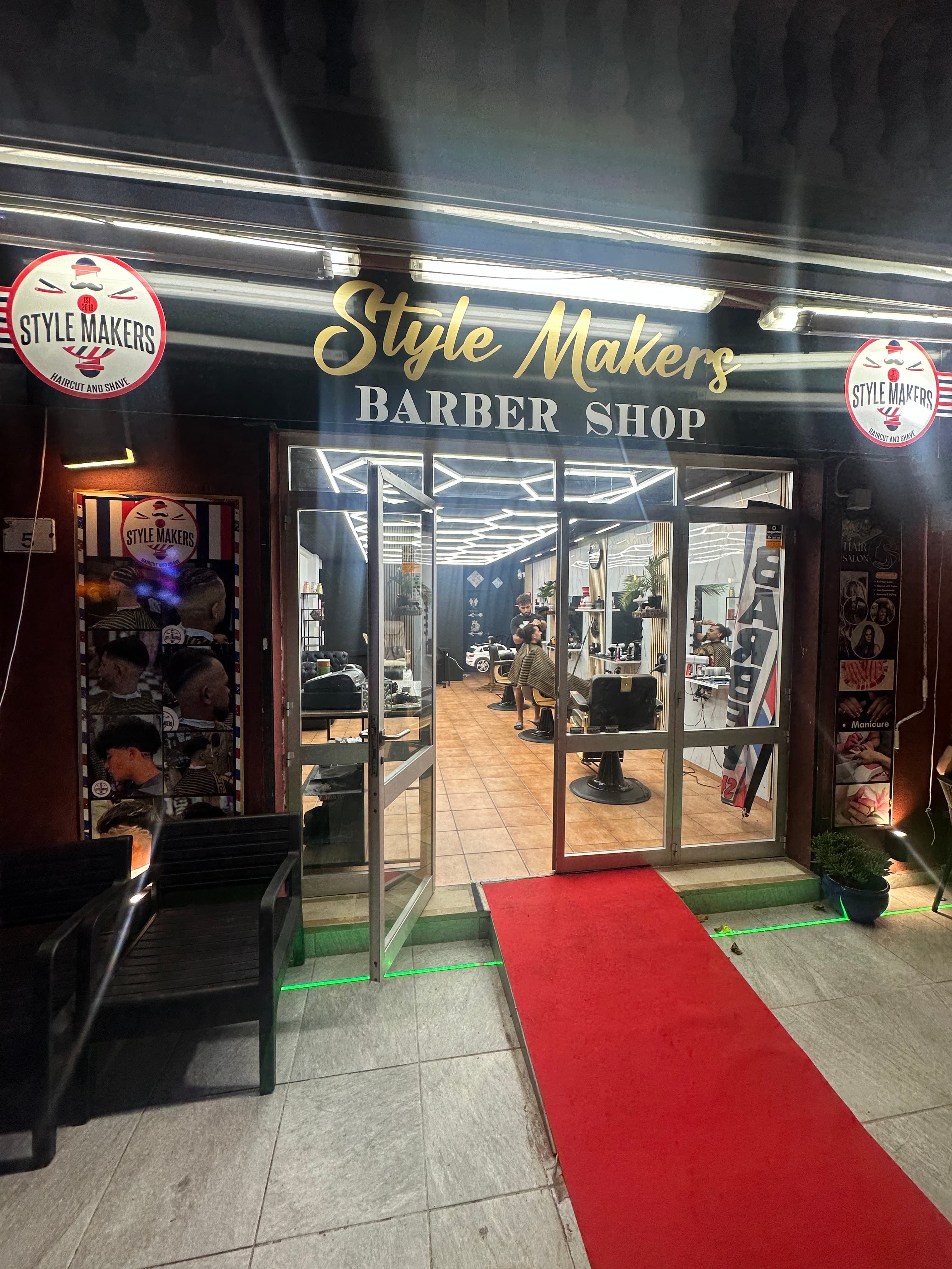 Fachada de Style Makers Barber Shop con alfombra roja y vista al interior iluminado.
